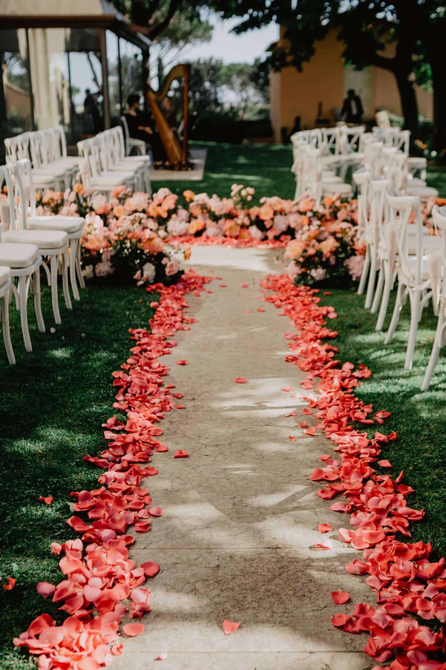 Aisle lined with rose petals runs between rows of white chairs in a garden.