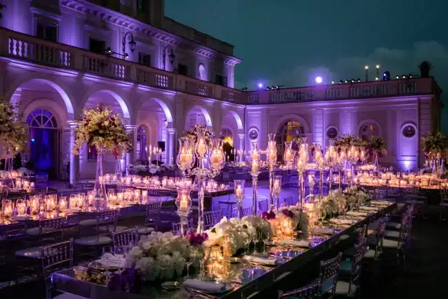 Wedding reception tables with candles and flowers are set outside a large, illuminated mansion at night.